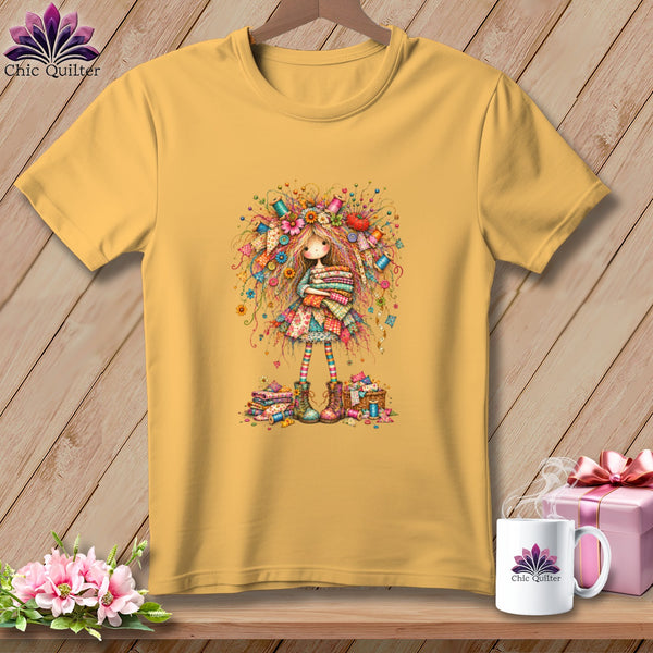 Gold t-shirt featuring a whimsical illustration of a girl holding folded fabric and surrounded by sewing notions.