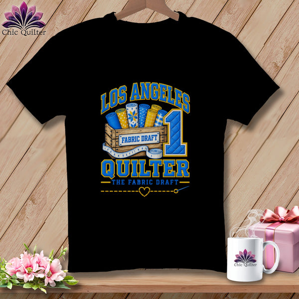 Black t-shirt featuring a Los Angeles Quilter graphic with a wooden crate of fabric rolls and a measuring tape.