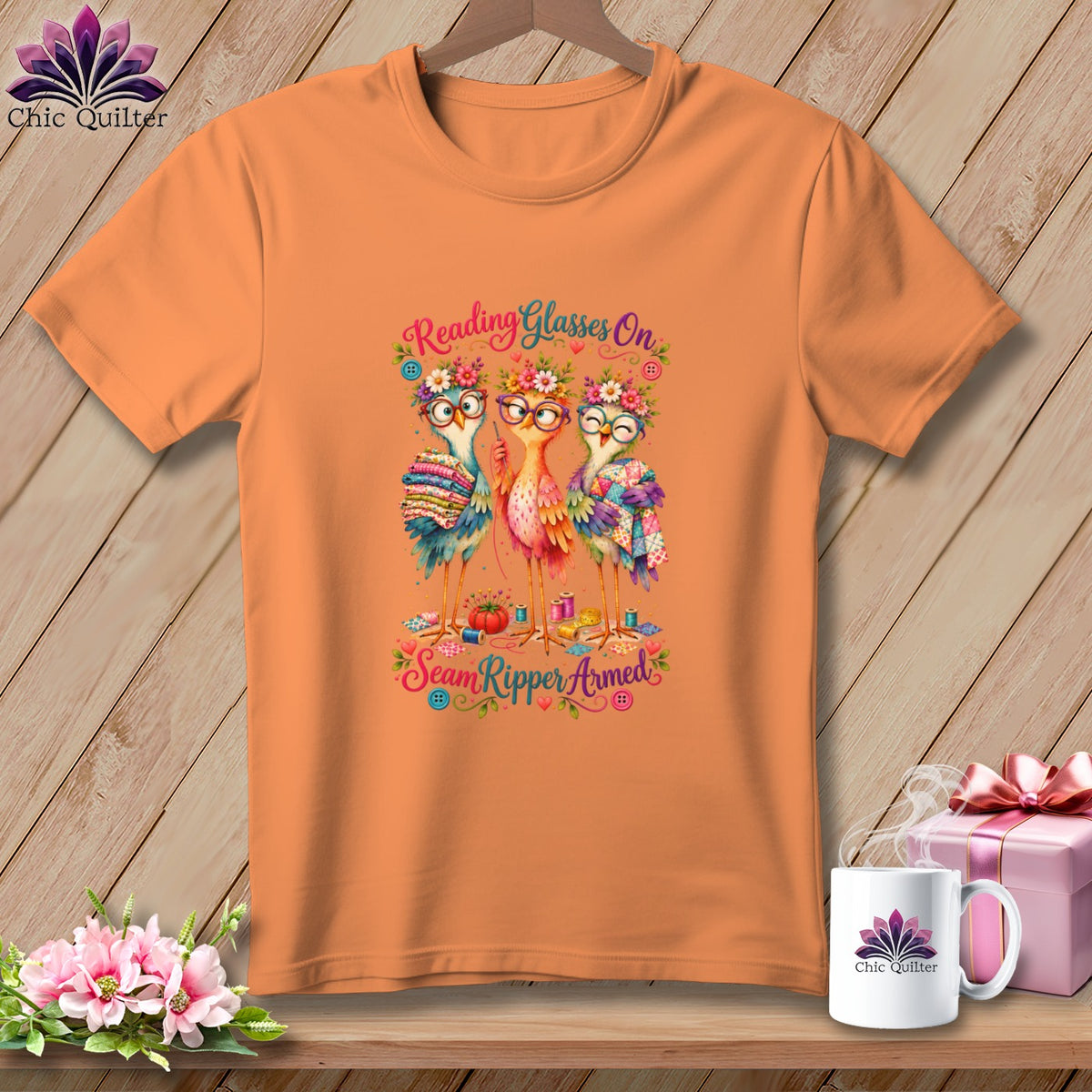 Orange t-shirt featuring three birds wearing glasses, floral wreaths, and holding sewing supplies with text.