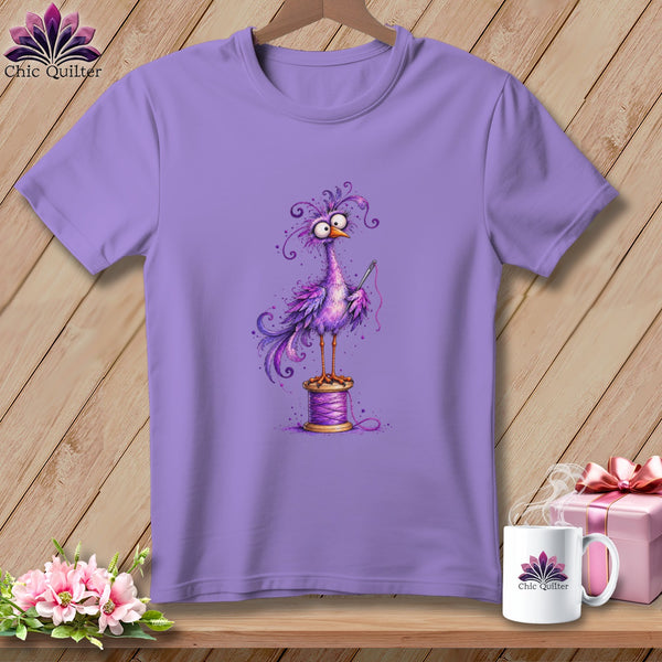 A purple short-sleeved t-shirt features a whimsical purple bird holding a needle while perched on a thread spool.