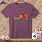 Purple t-shirt hanging on a wooden background, featuring a graphic of a woman riding a large spool of red thread.