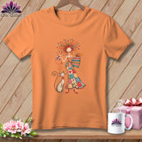 An orange short-sleeve t-shirt features a colorful illustration of a woman sewing alongside a cat.