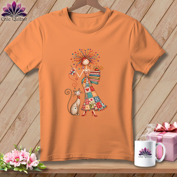 An orange short-sleeve t-shirt features a colorful illustration of a woman sewing alongside a cat.