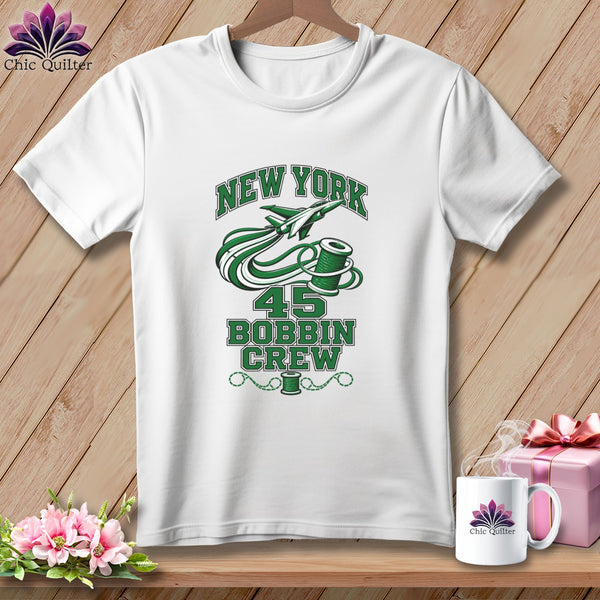 White t-shirt with a green graphic featuring a jet, a spool of thread, and text that reads New York 45 Bobbin Crew.