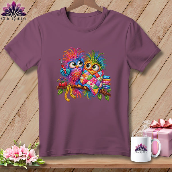 A purple t-shirt featuring a colorful graphic of two whimsical owls perched on a branch with sewing accessories.