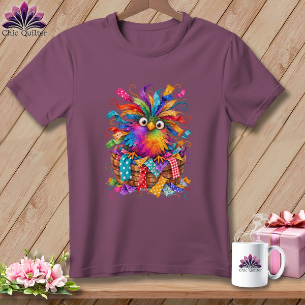 A purple t-shirt featuring a colorful, whimsical bird sitting in a basket of fabric scraps.