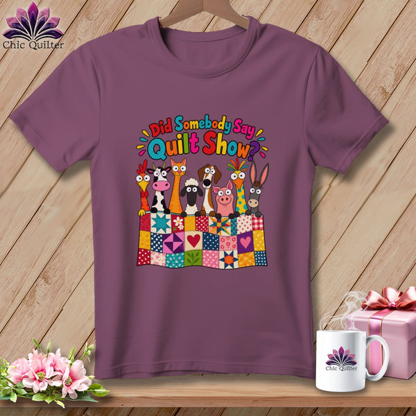 Purple t-shirt hanging on a wooden background, featuring cartoon farm animals behind a patchwork quilt and text reading "Did Somebody Say Quilt Show?".