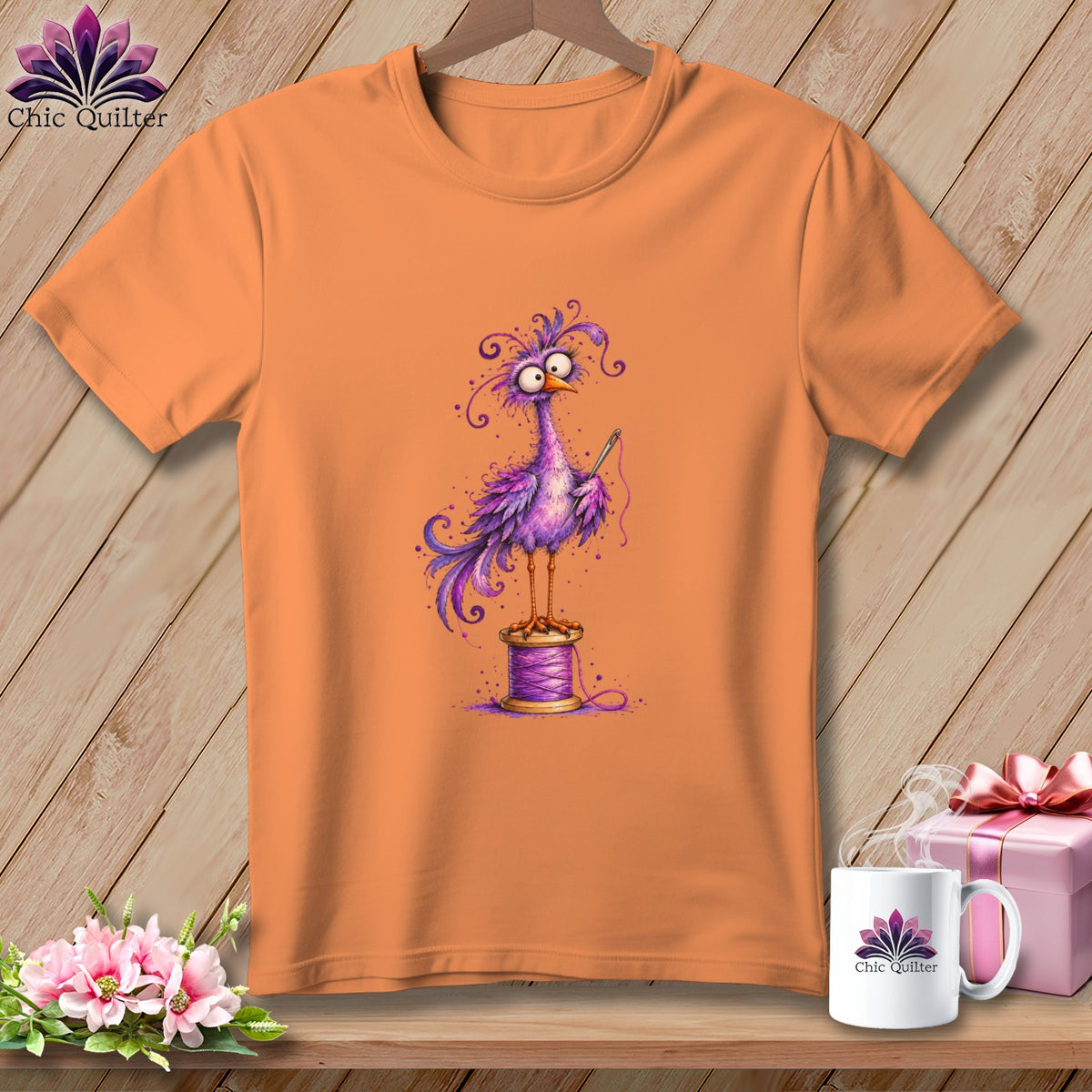 An orange t-shirt features a whimsical purple bird holding a needle while perched on a spool of thread.