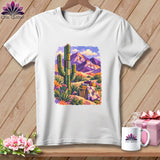 A white t-shirt hanging on a wooden background features a colorful graphic of a desert landscape with purple mountains and a saguaro cactus.