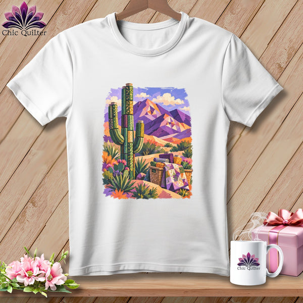 A white t-shirt hanging on a wooden background features a colorful graphic of a desert landscape with purple mountains and a saguaro cactus.