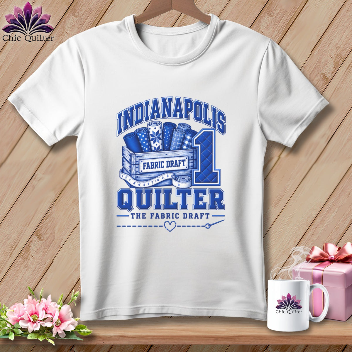 White t-shirt displaying a blue Indianapolis Quilter graphic with fabric rolls, a measuring tape, and a gift box.