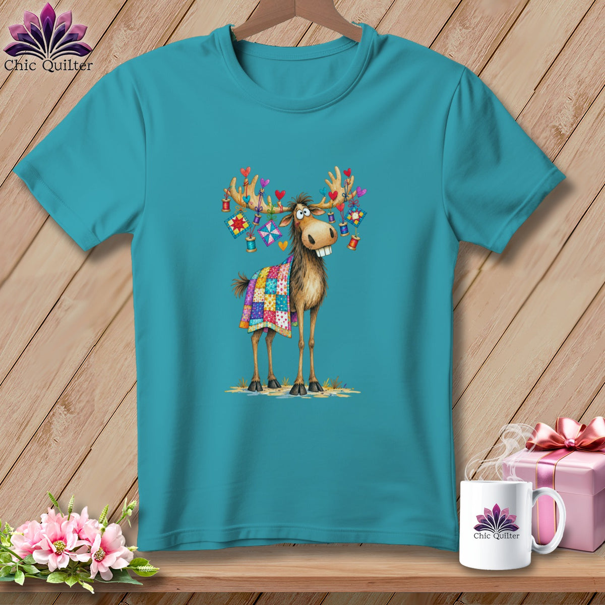 Teal t-shirt featuring a cartoon moose wearing a quilt and decorated antlers on a wooden background.