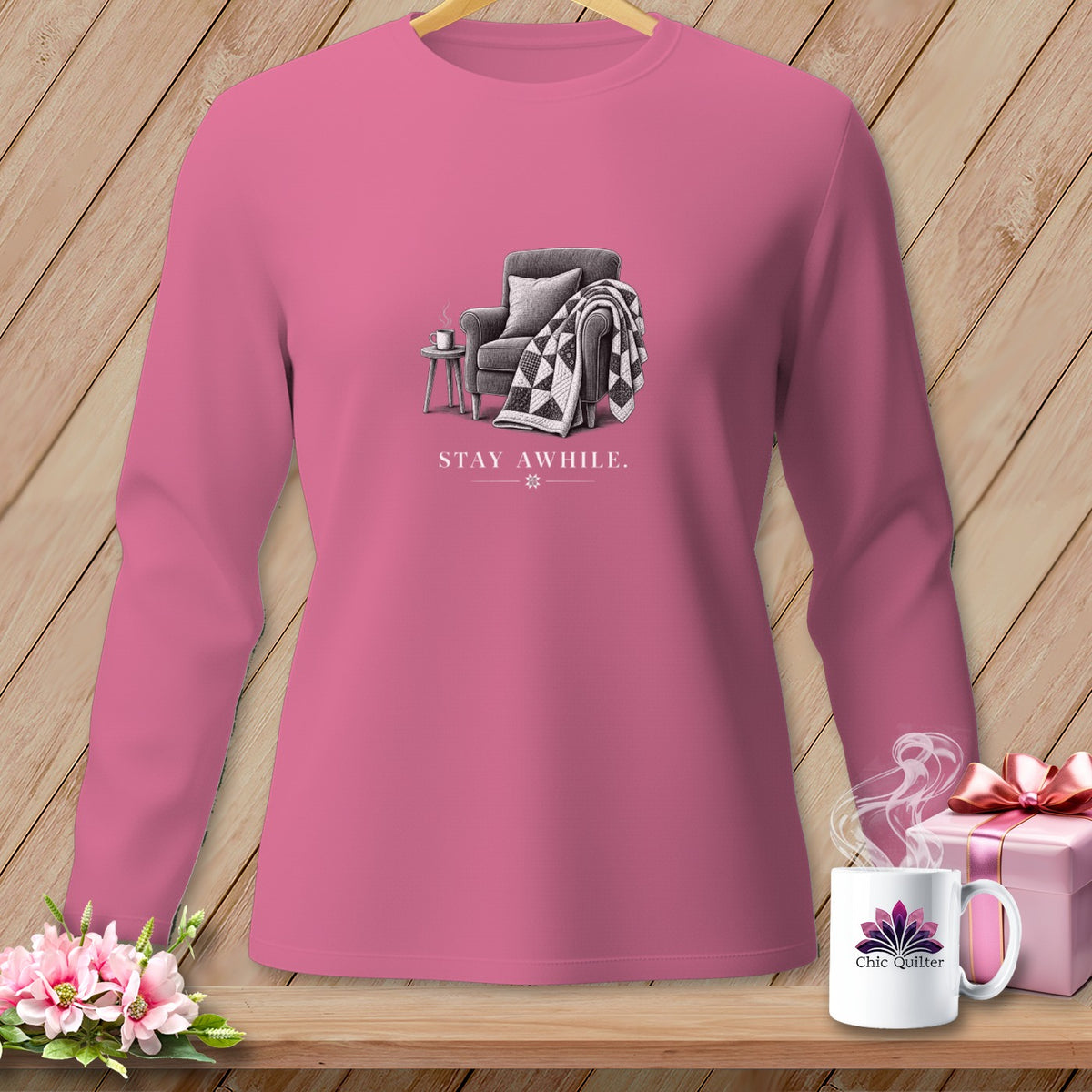 Pink long-sleeve shirt with a graphic of an armchair, quilt, and mug reading "STAY AWHILE."