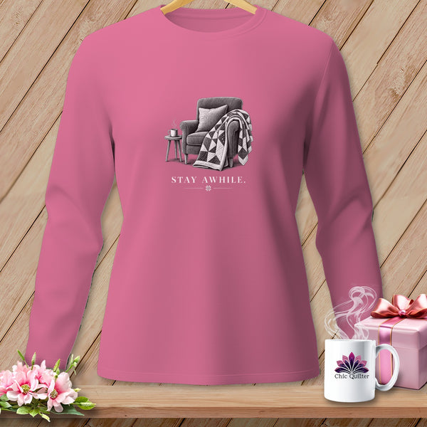 Pink long-sleeve shirt with a graphic of an armchair, quilt, and mug reading "STAY AWHILE."