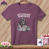 A plum-colored t-shirt on a wooden hanger with a graphic of a bear and raccoon quilting and the text 
