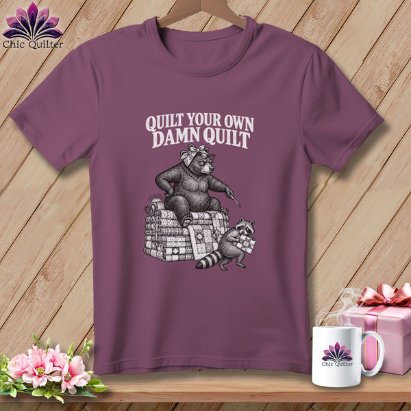 A plum-colored t-shirt on a wooden hanger with a graphic of a bear and raccoon quilting and the text "Quilt Your Own Damn Quilt".
