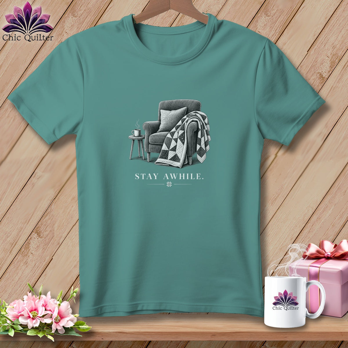 Teal t-shirt with a drawing of an armchair, a blanket, and a mug, and the text "STAY AWHILE."