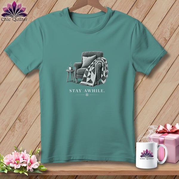 Teal t-shirt with a drawing of an armchair, a blanket, and a mug, and the text "STAY AWHILE."
