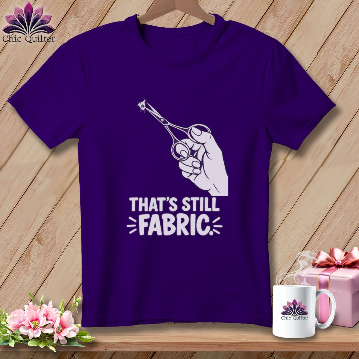 Purple t-shirt with graphic of hand holding scissors cutting fabric, text "That's Still Fabric".