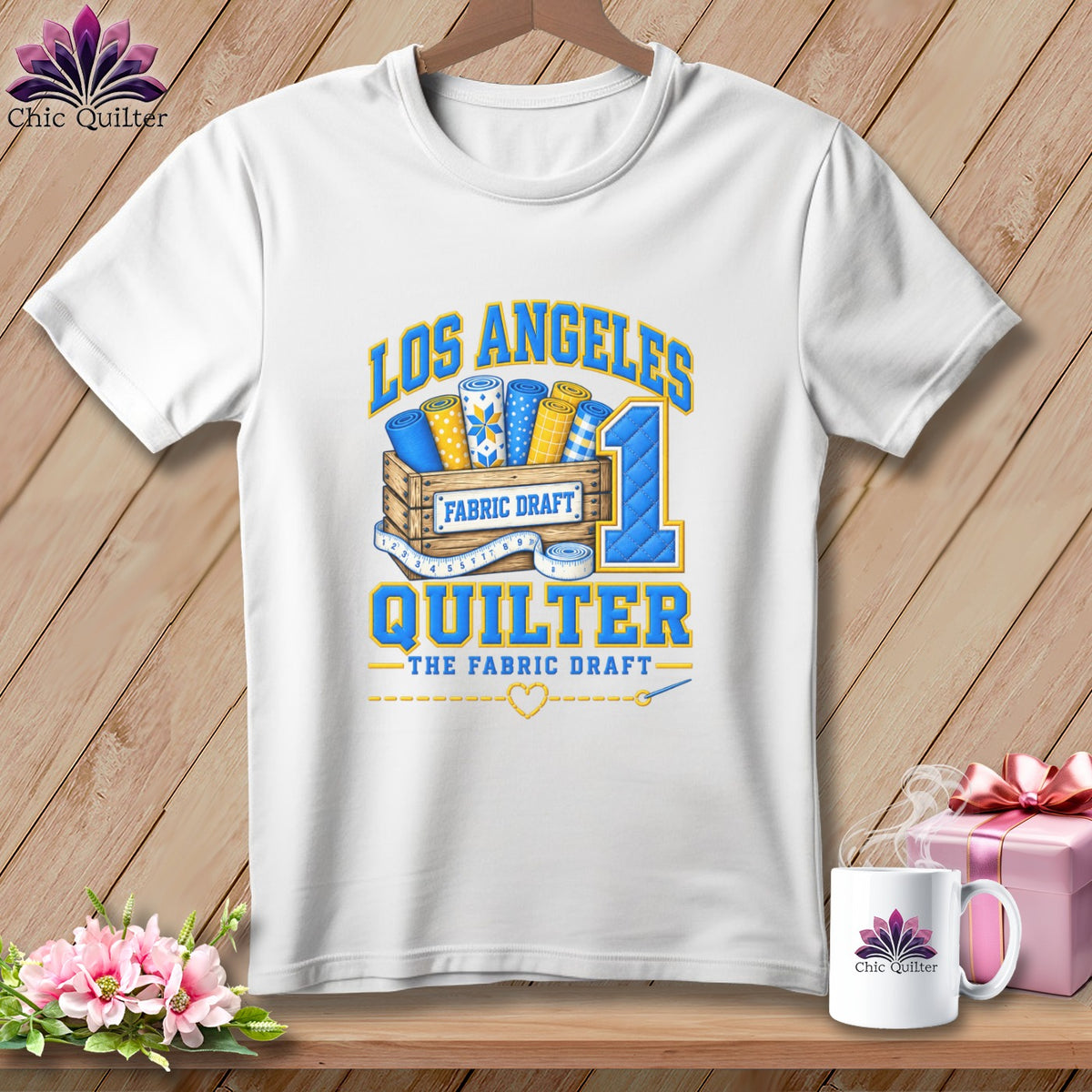 A white t-shirt hanging on a wooden background features a graphic with fabric rolls in a crate and text that reads Los Angeles Quilter.