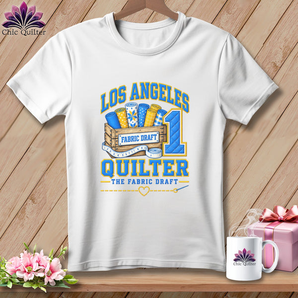 A white t-shirt hanging on a wooden background features a graphic with fabric rolls in a crate and text that reads Los Angeles Quilter.