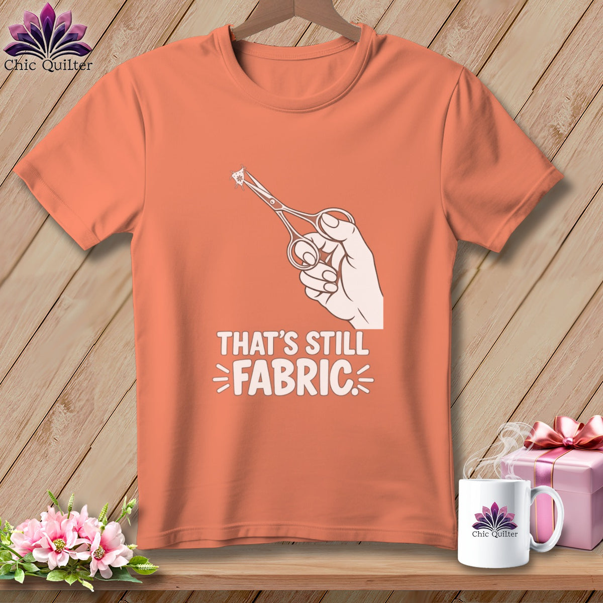 Peach t-shirt with a graphic of a hand holding scissors cutting a small piece of fabric, text reads "THAT'S STILL FABRIC".