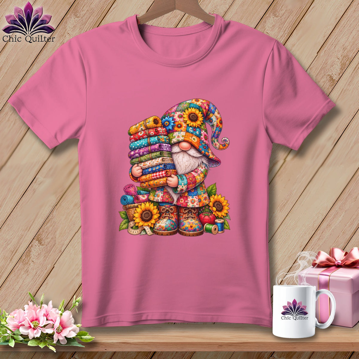 Pink t-shirt with a colorful gnome graphic holding a stack of fabric rolls, displayed on a wooden background.