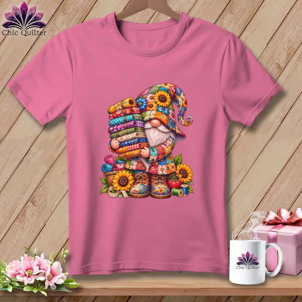 Pink t-shirt with a colorful gnome graphic holding a stack of fabric rolls, displayed on a wooden background.
