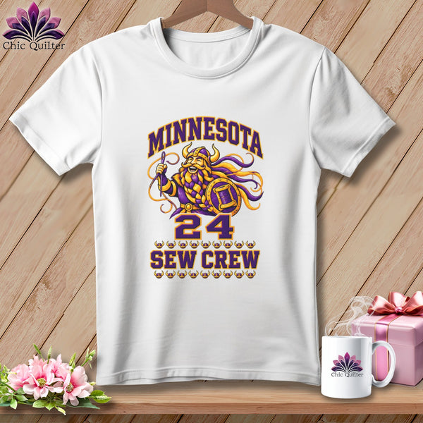A white t-shirt features a graphic with a cartoon Viking holding a needle, the text Minnesota Sew Crew 24, and matching mug and gift box on wood.
