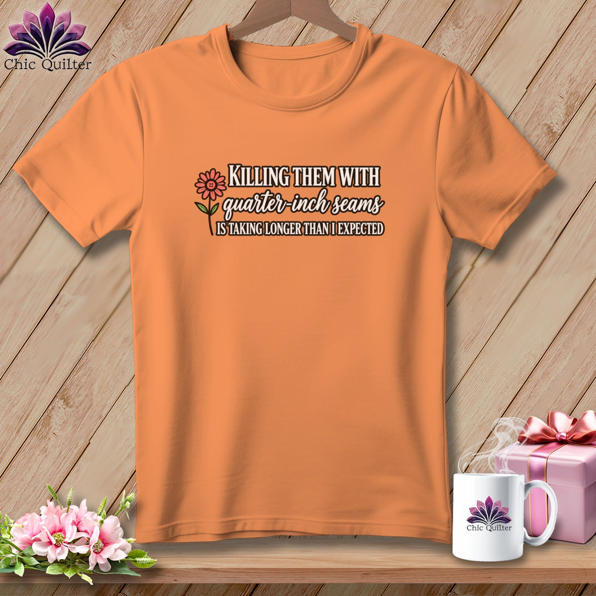 Orange t-shirt on a wooden background, featuring the text "KILLING THEM WITH quarter-inch seams IS TAKING LONGER THAN I EXPECTED" and a flower graphic.