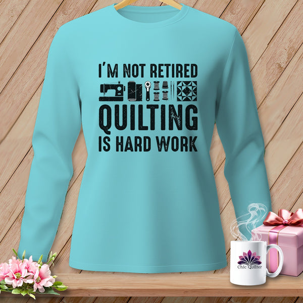 Light blue long-sleeve shirt with text "I'M NOT RETIRED QUILTING IS HARD WORK" and sewing graphics.