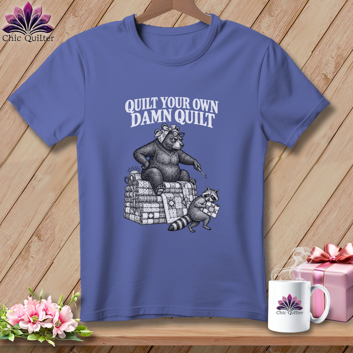 Purple t-shirt with a bear holding a needle sitting on a stack of quilts, and text saying Quilt Your Own Damn Quilt.