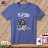Purple t-shirt with a bear holding a needle sitting on a stack of quilts, and text saying Quilt Your Own Damn Quilt.