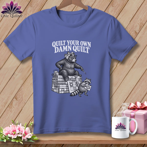 Purple t-shirt with a bear holding a needle sitting on a stack of quilts, and text saying Quilt Your Own Damn Quilt.