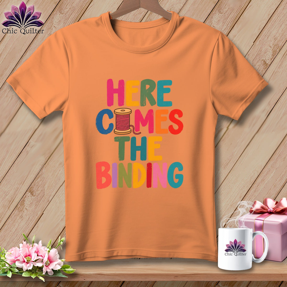 An orange t-shirt printed with colorful text reading Here Comes The Binding and a spool of pink thread.