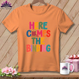 An orange t-shirt printed with colorful text reading Here Comes The Binding and a spool of pink thread.