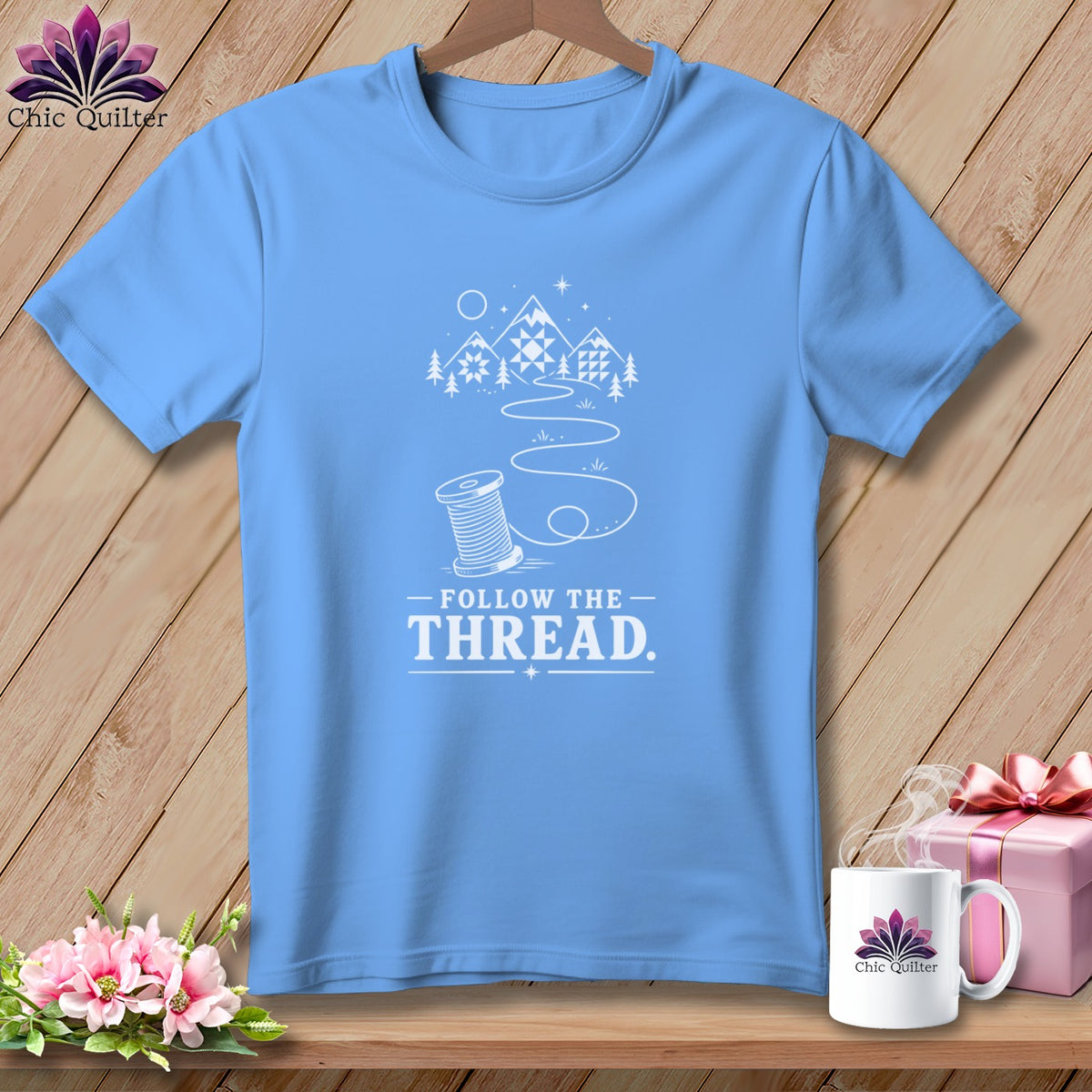 Light blue t-shirt with white graphic of a spool of thread leading to mountains and text "FOLLOW THE THREAD."