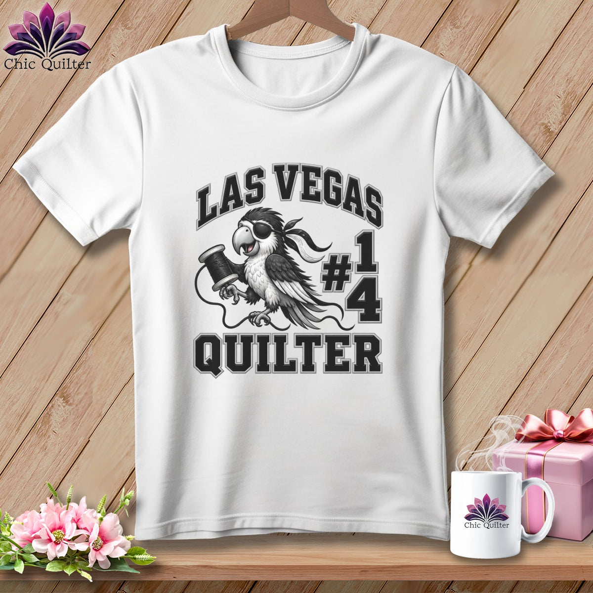 A white t-shirt hanging against a wooden background features a graphic of a parrot holding a spool of thread with the text "LAS VEGAS #1 QUILTER".