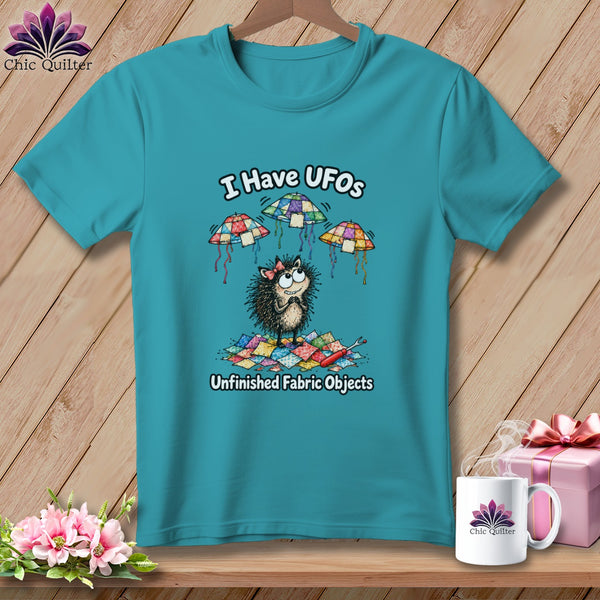 Teal t-shirt featuring a cartoon hedgehog, flying quilted UFO objects, and the text "I Have UFOs Unfinished Fabric Objects."