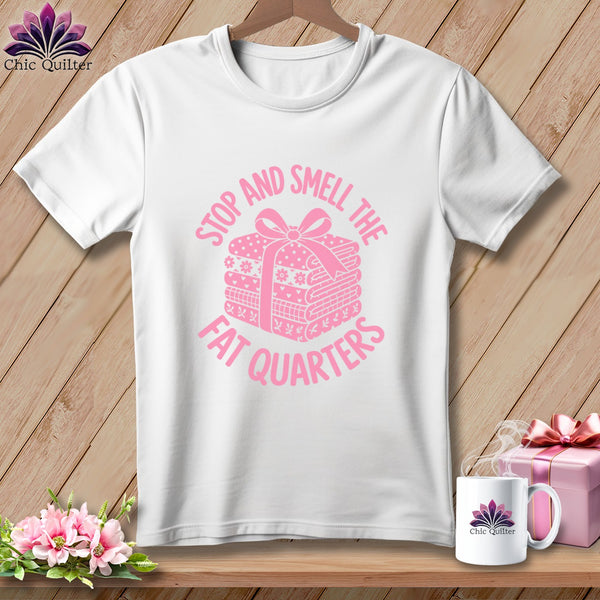 A white crewneck t-shirt featuring a pink graphic of a stack of folded fabric tied with a ribbon and the text Stop and Smell the Fat Quarters.