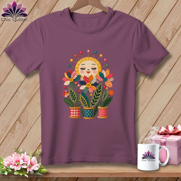 A mauve short-sleeve t-shirt features a colorful graphic of flower-filled thread spools topped with a smiling sun pin cushion.