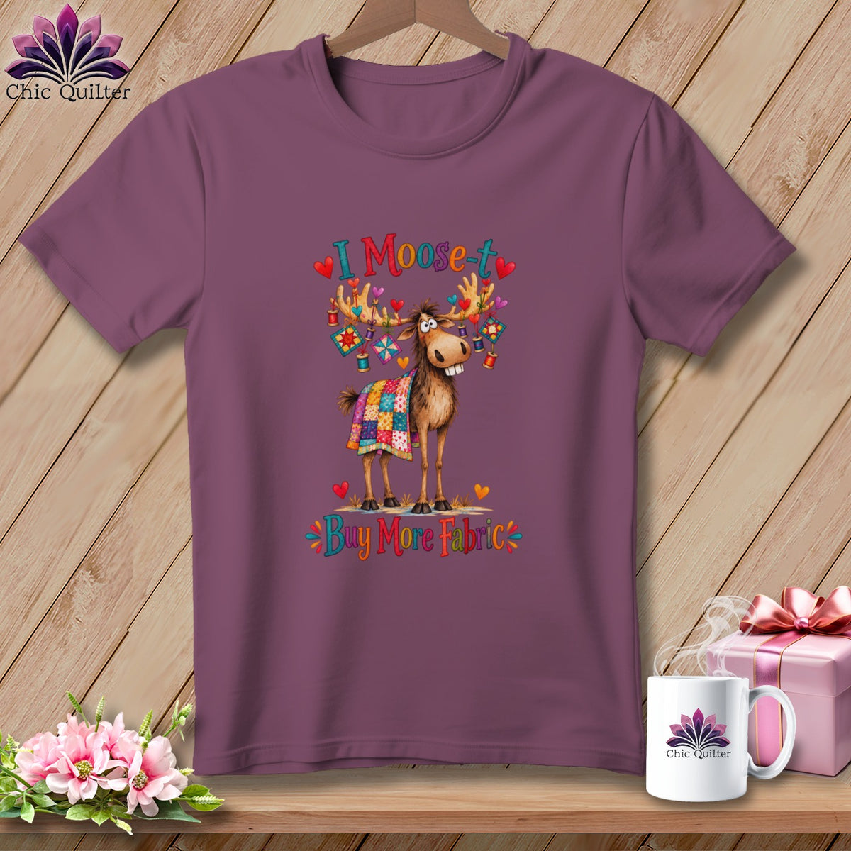 Purple t-shirt featuring a cartoon moose covered in a quilt, with text reading I Moose-t Buy More Fabric.