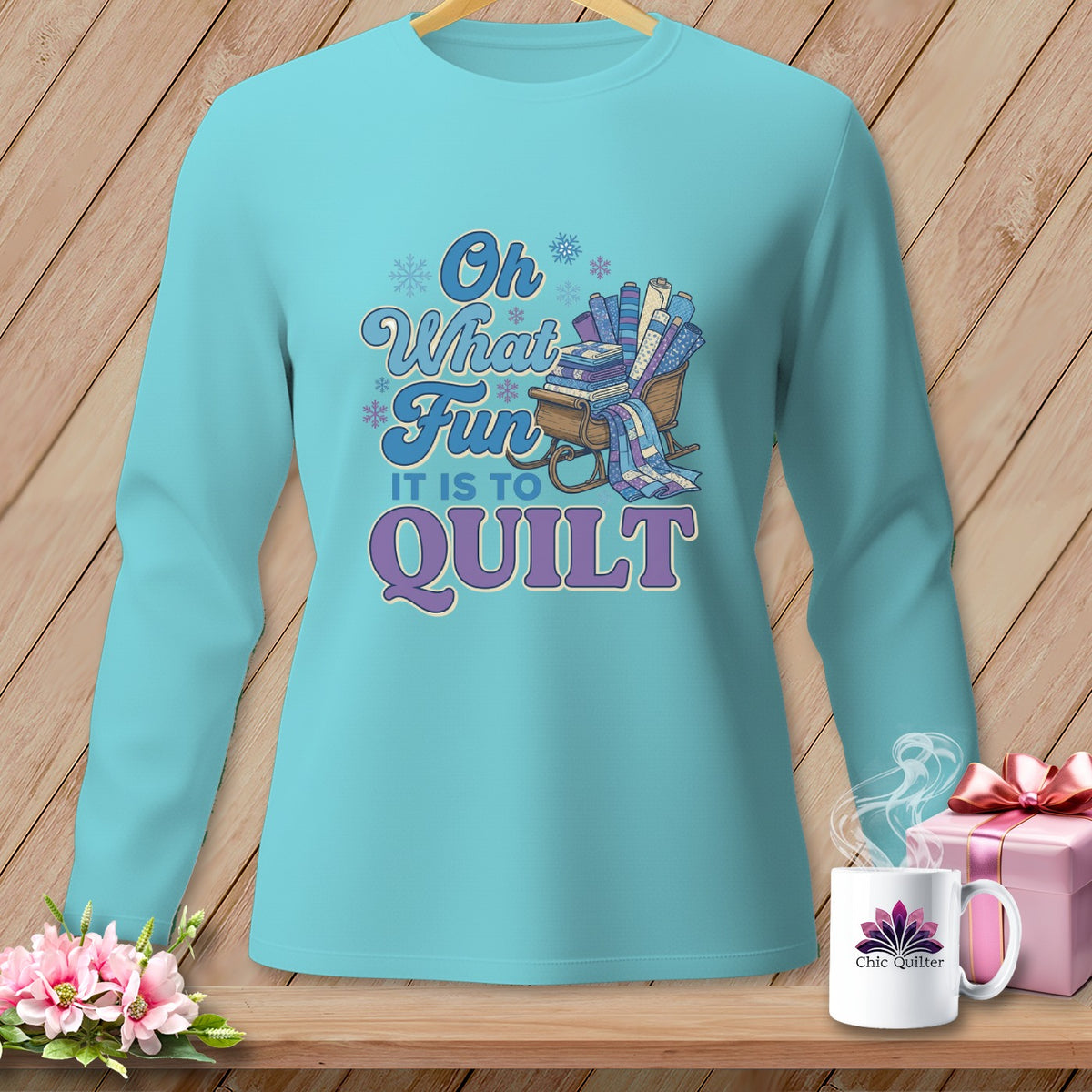Aqua long-sleeve shirt with "Oh What Fun It Is To Quilt" text and sled of fabric rolls.