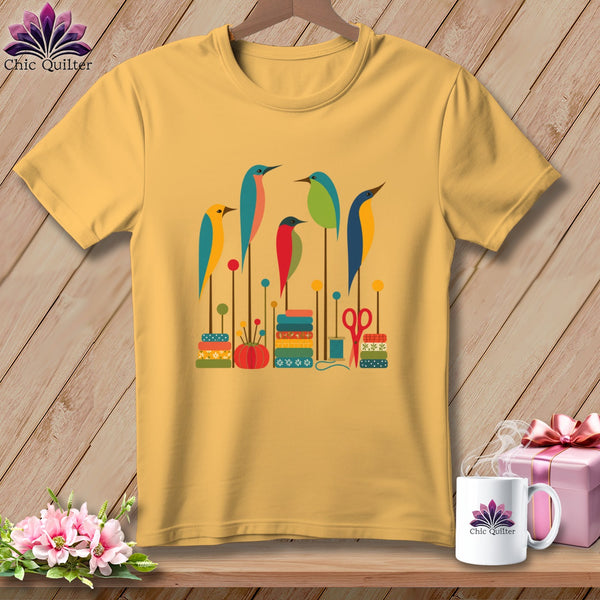 A yellow t-shirt features a graphic print of colorful stylized birds and sewing items on a wooden background.