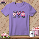 A light purple crewneck t-shirt with a graphic of a hand showing a peace sign wearing a thimble, a pink patchwork heart, and a stack of fabric with a spool of thread on top, with the text 