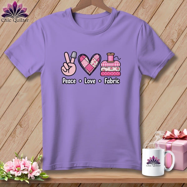 A light purple crewneck t-shirt with a graphic of a hand showing a peace sign wearing a thimble, a pink patchwork heart, and a stack of fabric with a spool of thread on top, with the text "Peace Love Fabric" below.