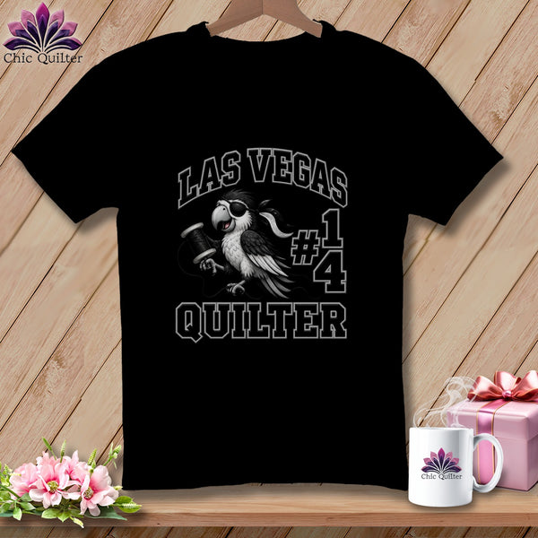 A black t-shirt hanging on a wooden background, featuring a graphic of a bird holding thread and text reading "Las Vegas #1 Quilter".