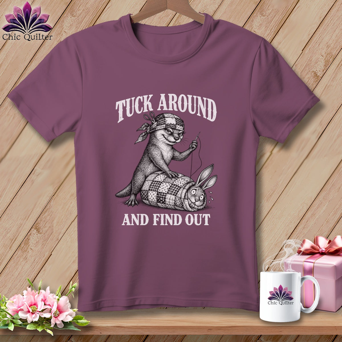 Mauve t-shirt on a wooden hanger with a graphic of an otter quilting a bunny and the text Tuck Around And Find Out.