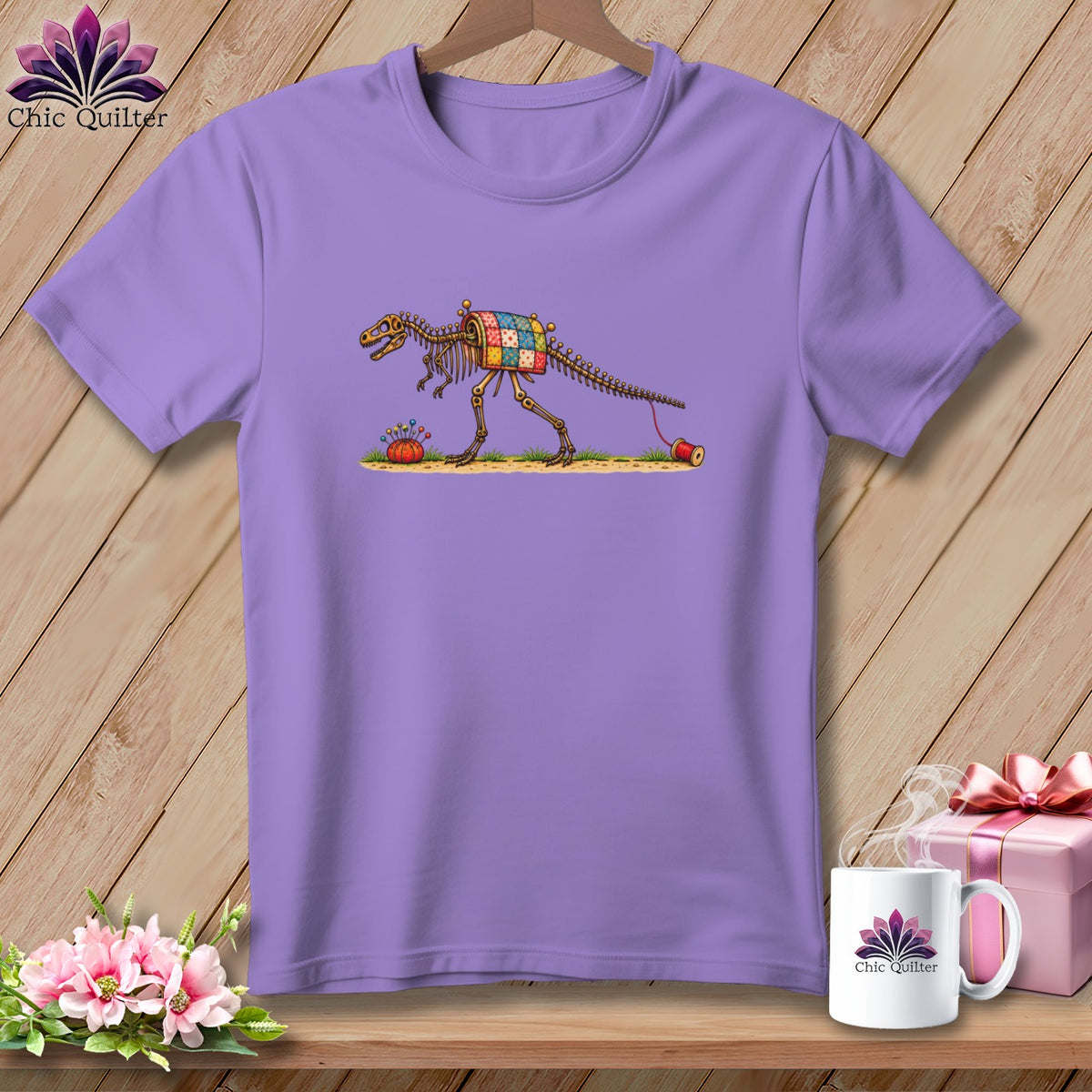 A light purple t-shirt featuring a dinosaur skeleton illustration wearing a colorful patchwork quilt.
