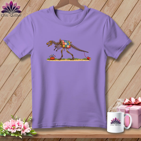 A light purple t-shirt featuring a dinosaur skeleton illustration wearing a colorful patchwork quilt.